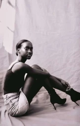 A Black woman seated on a grey textured backdrop wearing a dark sleeveless top, light denim shorts, and black high-heeled boots.