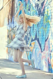 Young girl in plaid dress twirling outdoors on a sidewalk against a colourful graffiti wall background