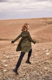 Woman in an olive green coat walking across a dry grassy meadow under an overcast sky, captured by Brian Sassmann.