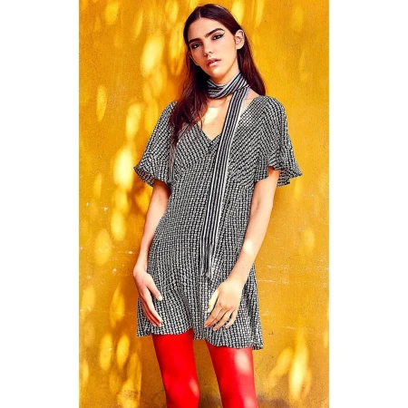 Model in patterned black and white dress and scarf wearing red trousers against a mottled yellow wall