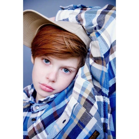 Close-up portrait of a child with red hair and blue eyes wearing a blue plaid shirt and a tan cap by Brian Sassmann.