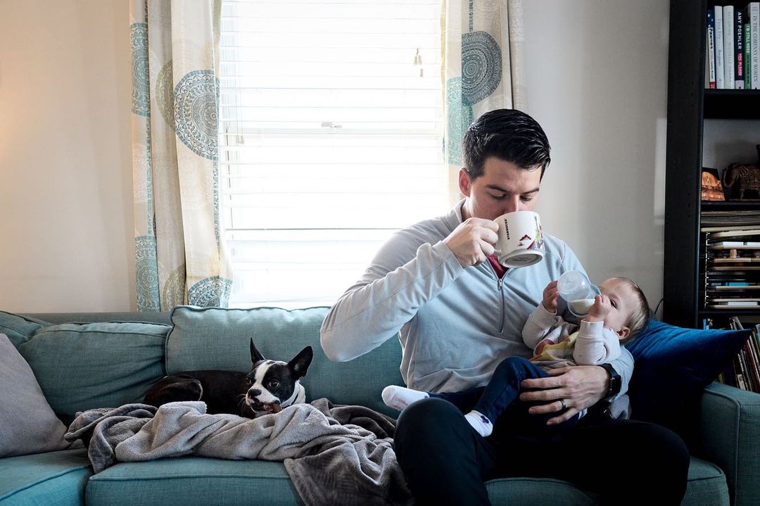 Man drinking coffee while feeding baby a bottle on a teal couch with a Boston terrier dog; indoor home setting near a window.