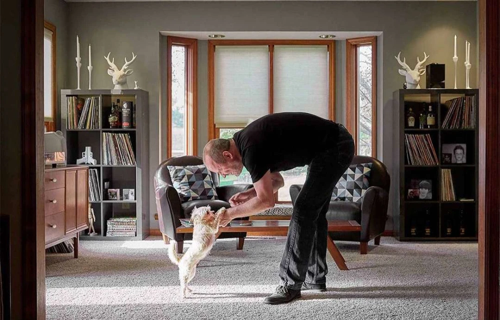 Adult male interacts with a small dog indoors in a home setting with bookshelves, windows, and interior decor, creating a cozy atmosphere.