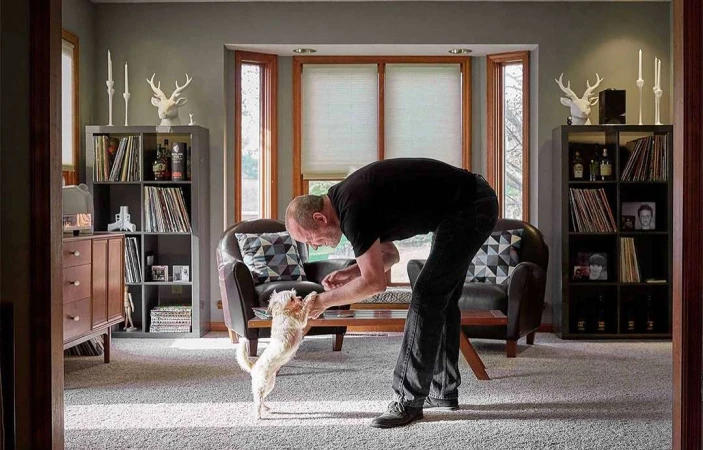 Adult male interacts with a small dog indoors in a home setting with bookshelves, windows, and interior decor, creating a cozy atmosphere.