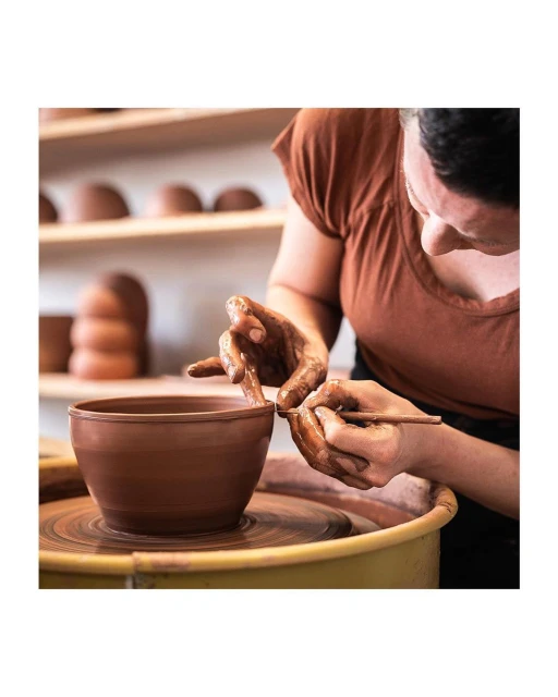 Adult female potter shapes wet clay bowl on a spinning pottery wheel in her art studio; a handmade ceramic pottery display is in the background.