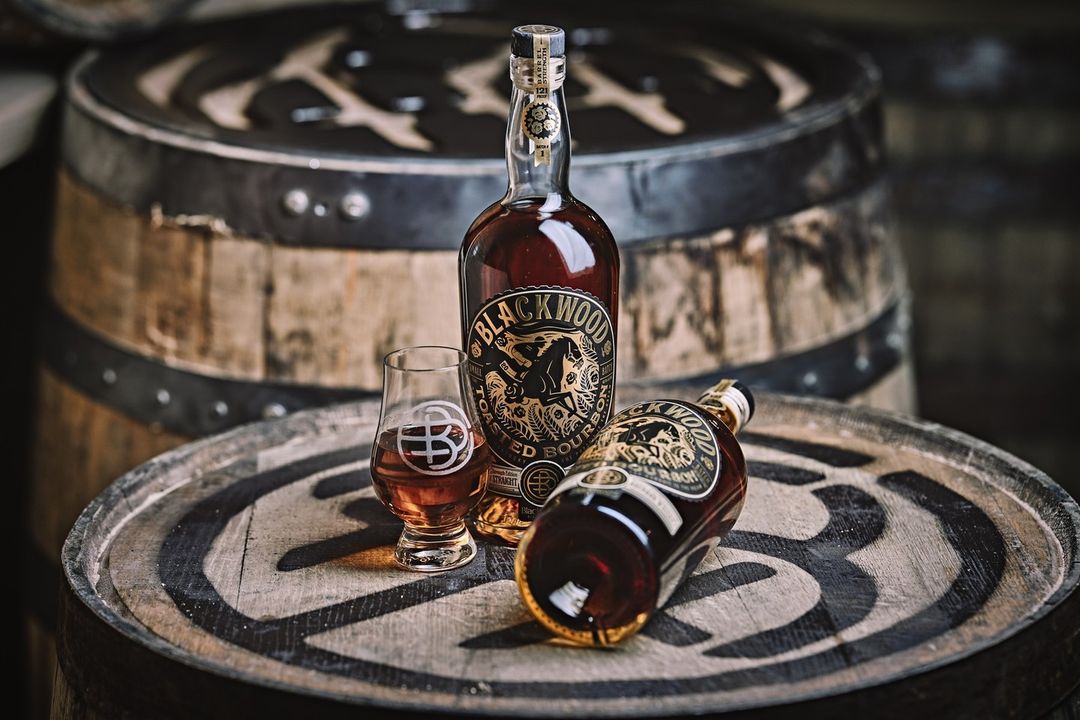 Two Blackwood Toasted Bourbon bottles and a glass sit atop a barrel with blurred barrels in the background. 