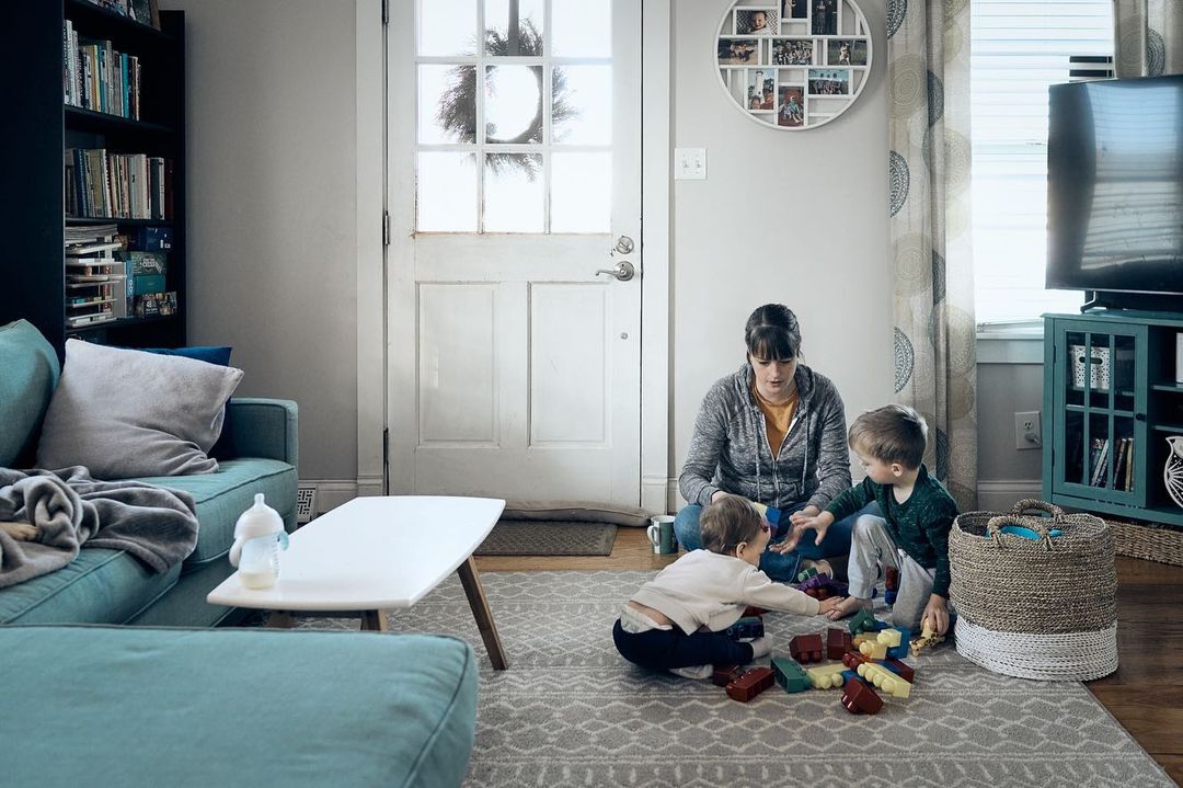 A mother plays with her two young children on the floor of a bright living room, surrounded by toys, furniture and interior decorations.
