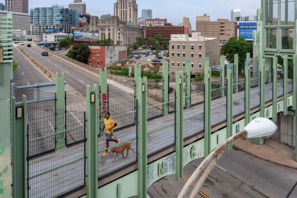 Adult male jogs with his dog on an enclosed pedestrian bridge with the city skyline and highway in the background on an overcast day.
