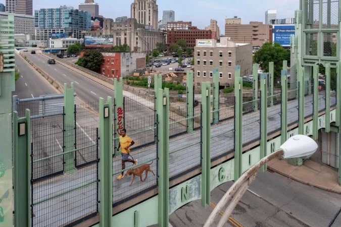 Adult male jogs with his dog on an enclosed pedestrian bridge with the city skyline and highway in the background on an overcast day.