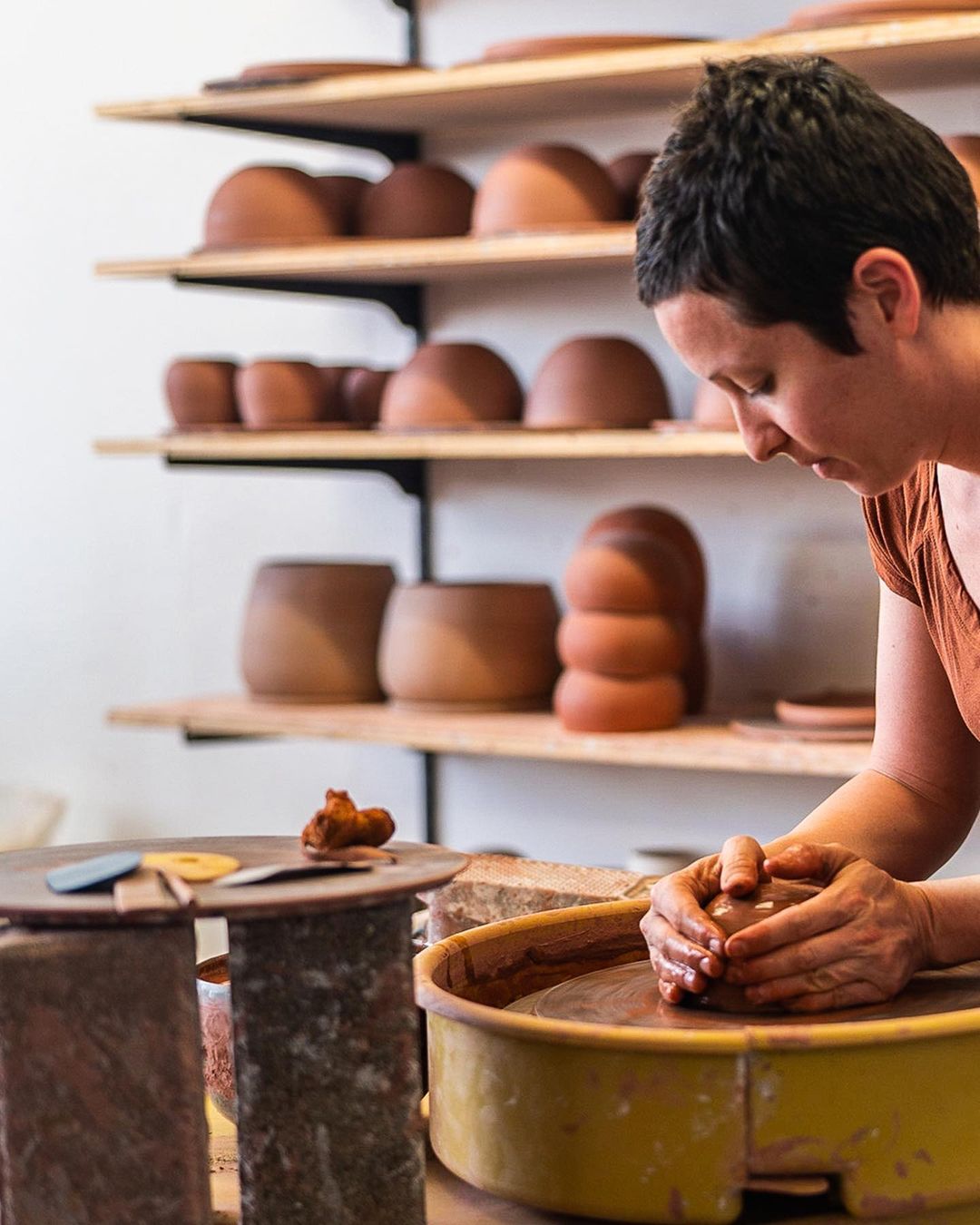 Woman shaping clay on a pottery wheel in a workshop. Shelves display finished clay pieces in the background in a neutral-toned studio setting.