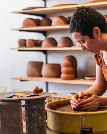 Woman shaping clay on a pottery wheel in a workshop. Shelves display finished clay pieces in the background in a neutral-toned studio setting.