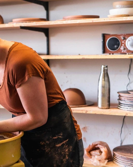 Adult female potter wearing an apron at a pottery wheel in a studio with shelves of clay plates and dishes, and equipment.