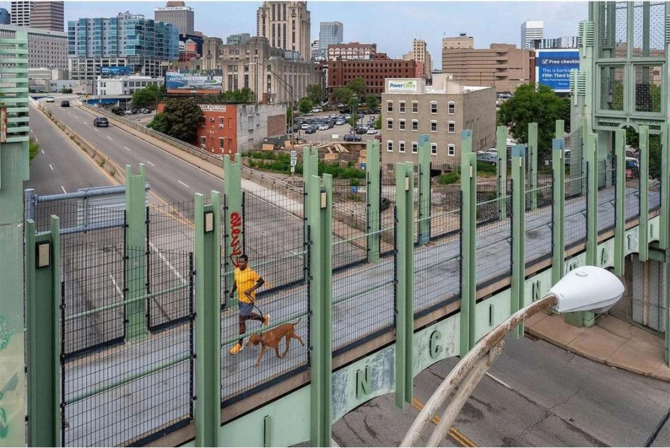 A man runs with his dog on a bridge in a city. Buildings are visible in the background and the image is taken from above.
