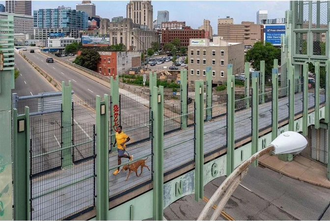 A man runs with his dog on a bridge in a city. Buildings are visible in the background and the image is taken from above.