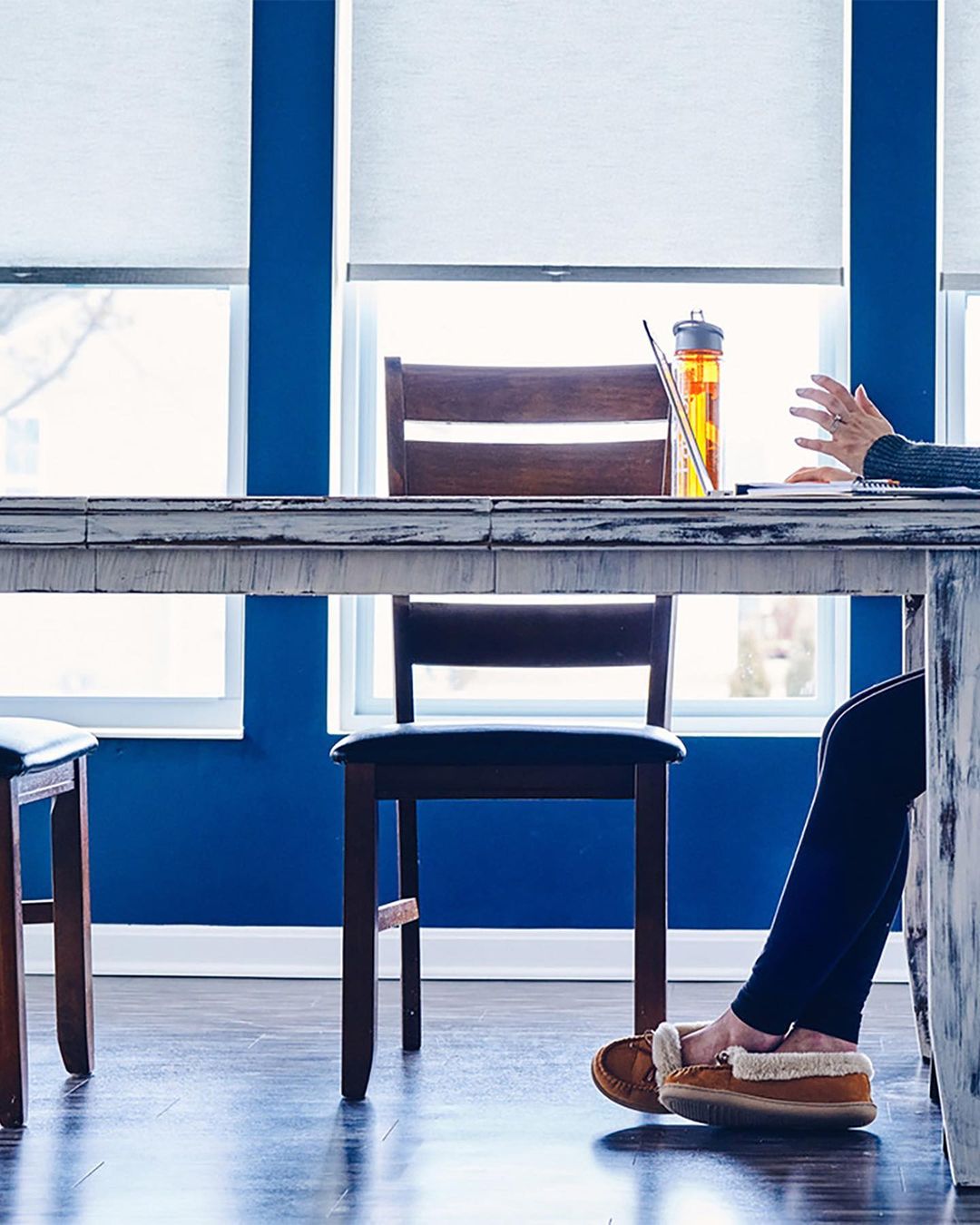 Person wearing moccasins sits at a wooden table with a laptop and a water bottle in a blue room with natural light from window.
