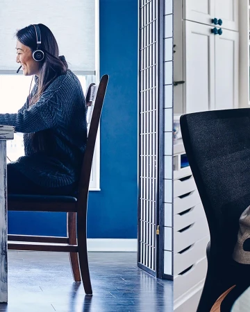 Woman working from home at a white table, wearing a headset in a blue-walled room, with a screen divider, and white cabinets.