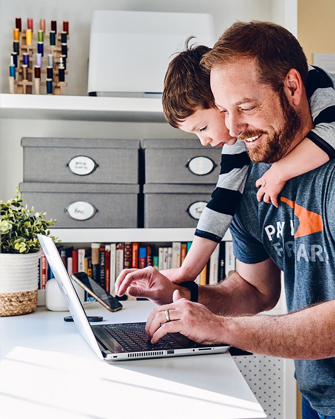 A man and a child are at a desk; the man is working on a laptop while the child stands behind him with his arms around his neck.