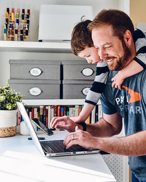 A man and a child are at a desk; the man is working on a laptop while the child stands behind him with his arms around his neck.