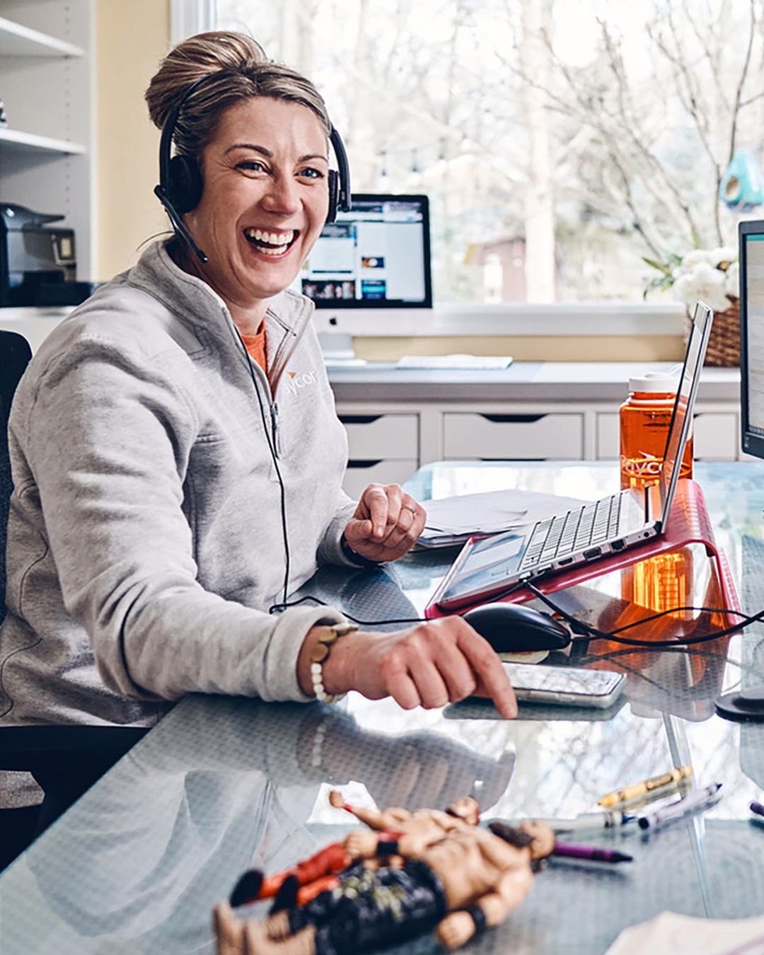 Woman with headset smiling and working at her home office desk with laptop, monitor, glass top desk, and toy figures.