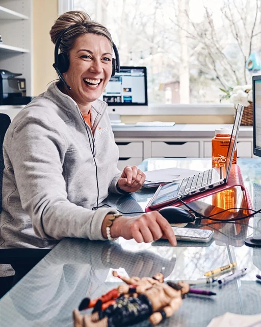 Woman with headset smiling and working at her home office desk with laptop, monitor, glass top desk, and toy figures.