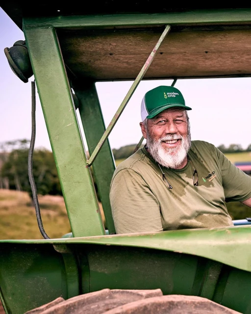 Smiling elderly man with beard and hat sits inside the cab of a vintage green tractor, with fields and sky visible in the background.