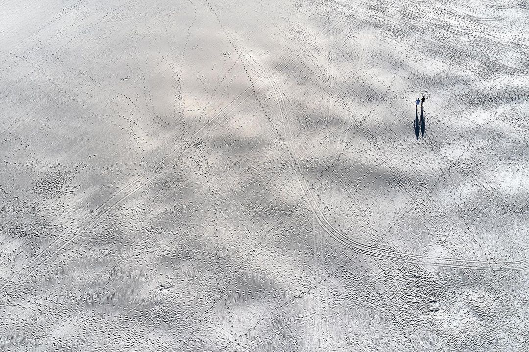 Overhead shot of two people walking on a sandy beach with light grey tones, leaving behind trails and shadows on the expansive surface.