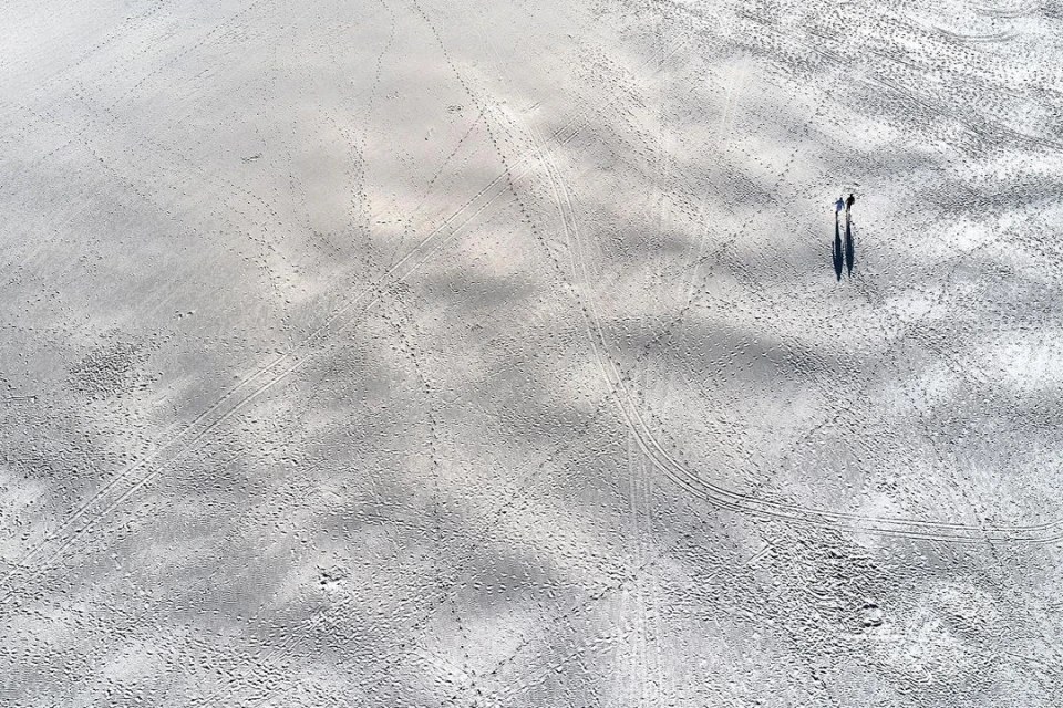 Overhead shot of two people walking on a sandy beach with light grey tones, leaving behind trails and shadows on the expansive surface.