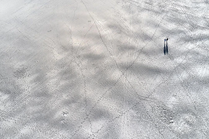 Overhead shot of two people walking on a sandy beach with light grey tones, leaving behind trails and shadows on the expansive surface.