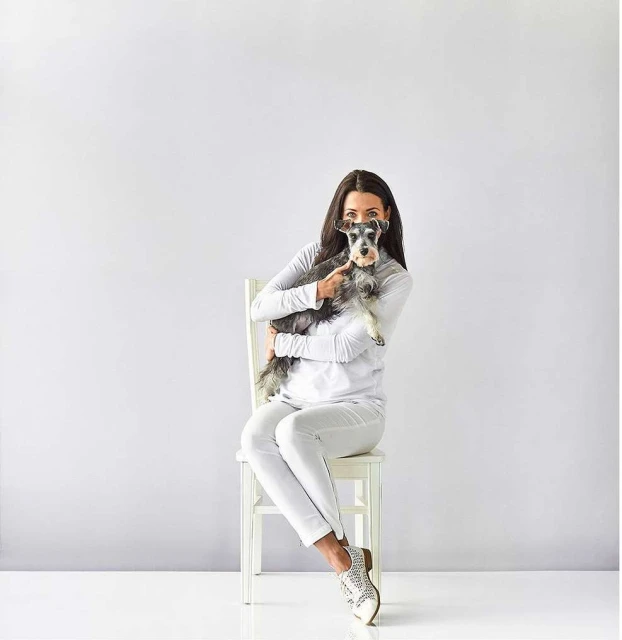 Adult woman sits on a chair in a photo studio, holding a Schnauzer dog in her arms. The background is plain white.