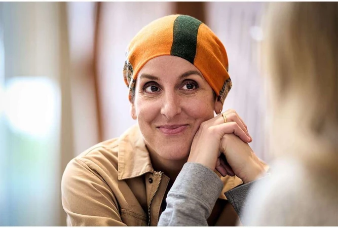 Close-up of a smiling adult woman wearing an orange and green head covering and a tan jacket indoors with a blurred background.