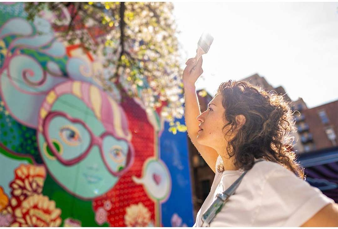 A woman stands outdoors on a sunny day painting a colorful mural on a building wall, holding a brush up to the artwork.