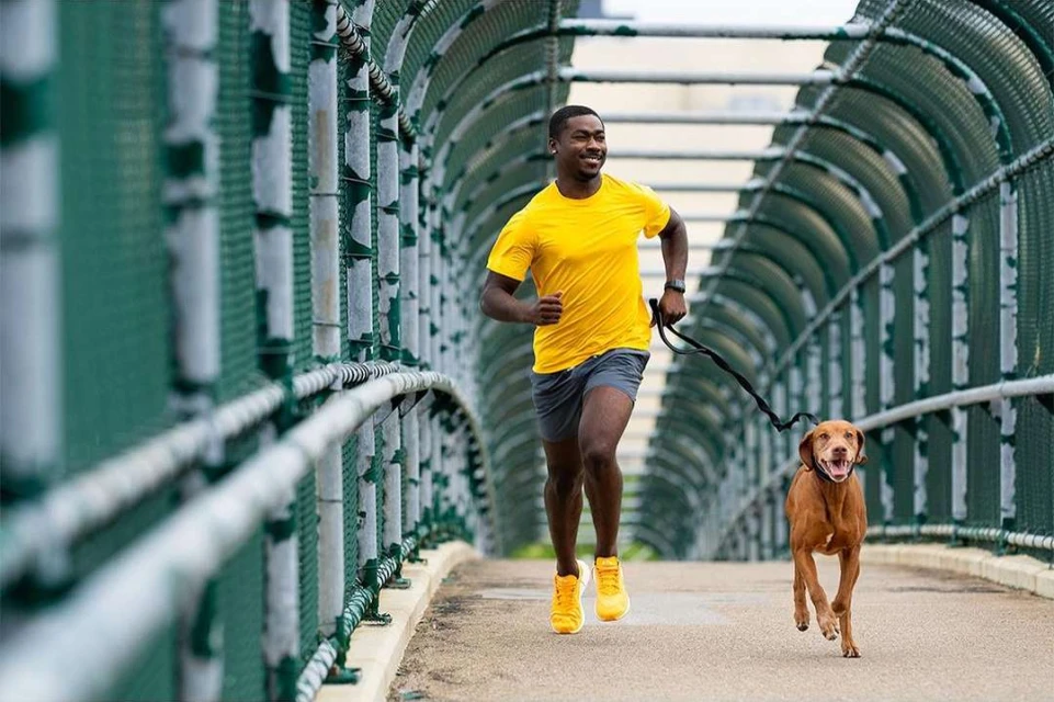 An African American man runs with his brown dog on a walkway with green metal arched fencing, wearing a yellow shirt and grey shorts.