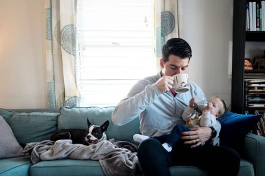 Man drinking from mug while holding baby with bottle on a teal sofa with a small dog
