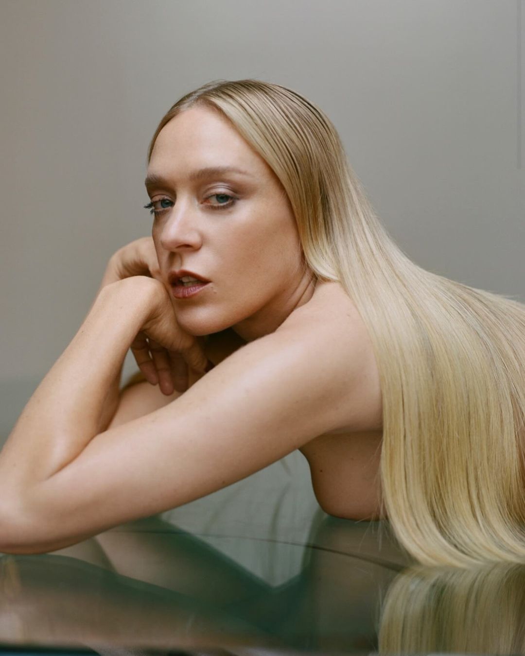 Blonde adult female portrait with hands near face and long hair, lying on a smooth, reflective surface against a plain background.