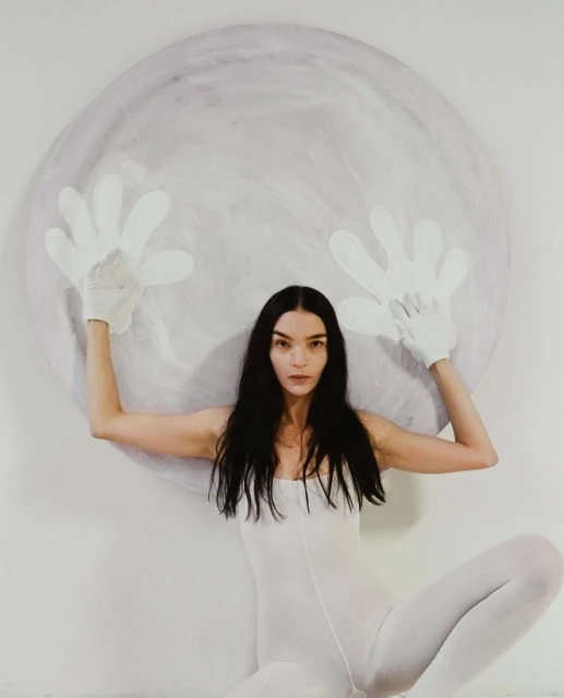 Woman in white bodysuit poses with large round prop art piece wearing oversized white hand gloves against a white background in studio.