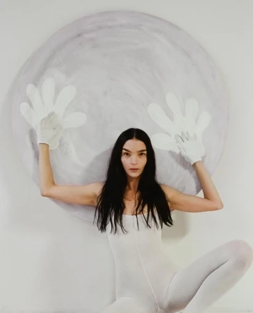 Woman in white bodysuit poses with large round prop art piece wearing oversized white hand gloves against a white background in studio.