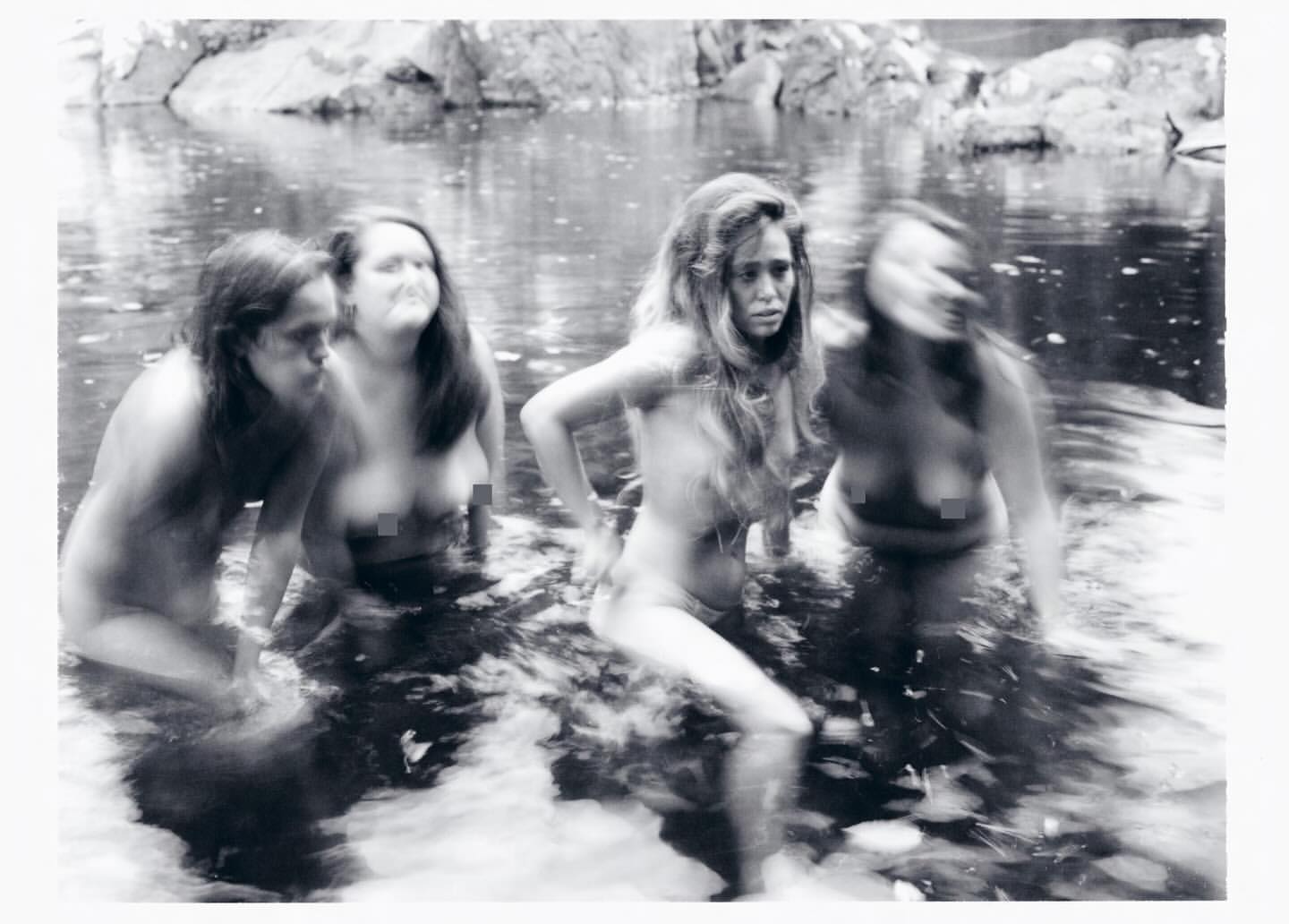 Monochrome photograph of four women wading in a shallow river surrounded by rocks and dense foliage, captured with motion blur.