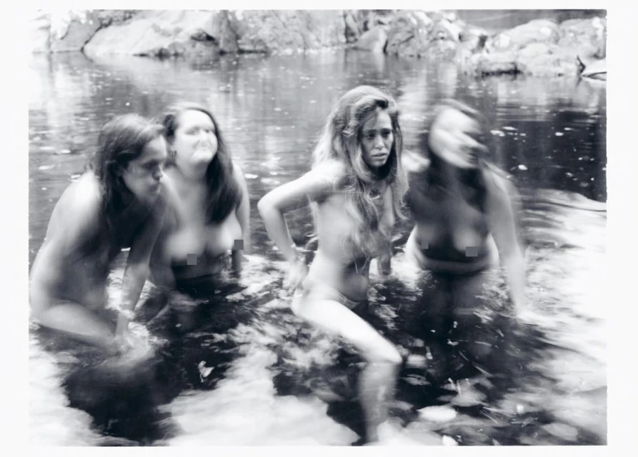 Monochrome photograph of four women wading in a shallow river surrounded by rocks and dense foliage, captured with motion blur.