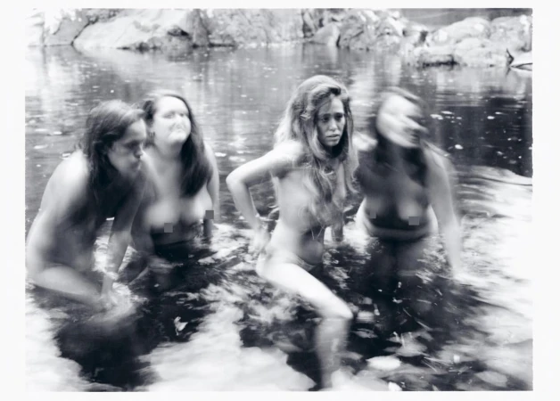 Monochrome photograph of four women wading in a shallow river surrounded by rocks and dense foliage, captured with motion blur.