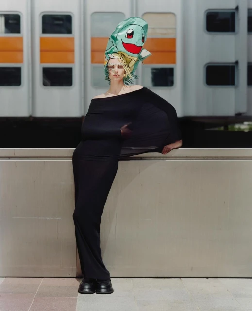 Woman in black dress and Squirtle balloon headpiece stands by grey wall with blurry train background in a fashion-style portrait shot.