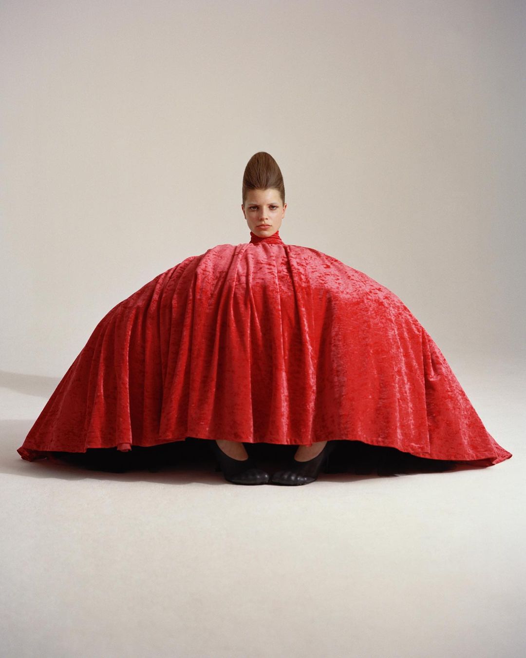 Fashion shot of a person in a draped red velvet fabric garment with a tall beehive hairstyle, set against a plain light backdrop.