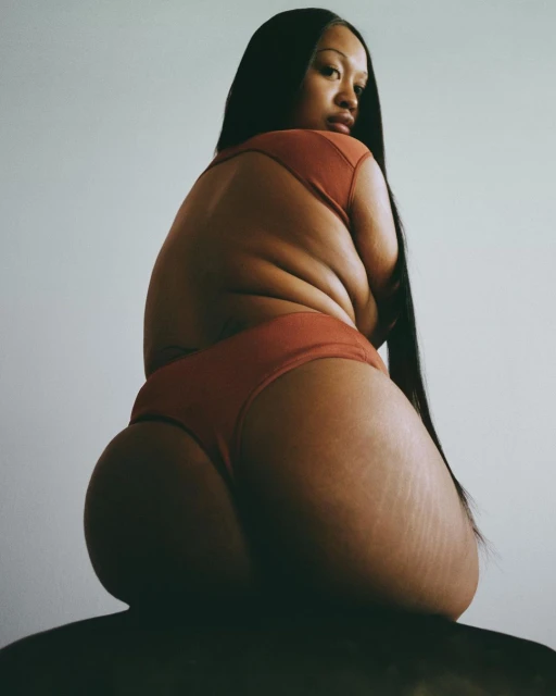 Portrait of a plus-size woman sitting with her back to the camera, wearing a terracotta-colored top and bottom, long black hair, indoors.