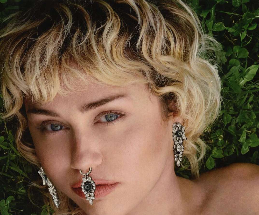 Close-up of a blonde woman with blue eyes and ornamental piercings laying on a green clover lawn, wearing jeweled earrings.