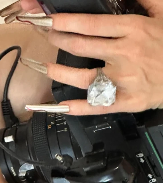 Close-up of a person's hands with long, silver, sculpted nails, wearing a large ring, and holding a camera, focusing on fashion and style.