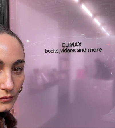 Adult woman stands near window with 'CLIMAX books, videos and more' text. Indoor setting with commercial display visible through glass.