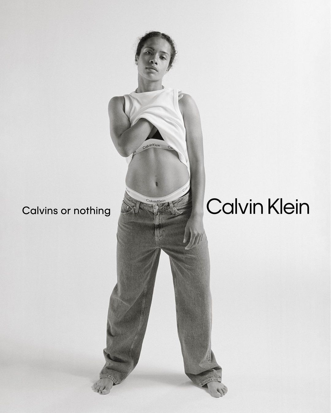 Black and white portrait of a woman lifting her white t-shirt wearing denim jeans and branded underwear against a plain background
