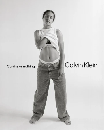 Black and white portrait of a woman lifting her white t-shirt wearing denim jeans and branded underwear against a plain background
