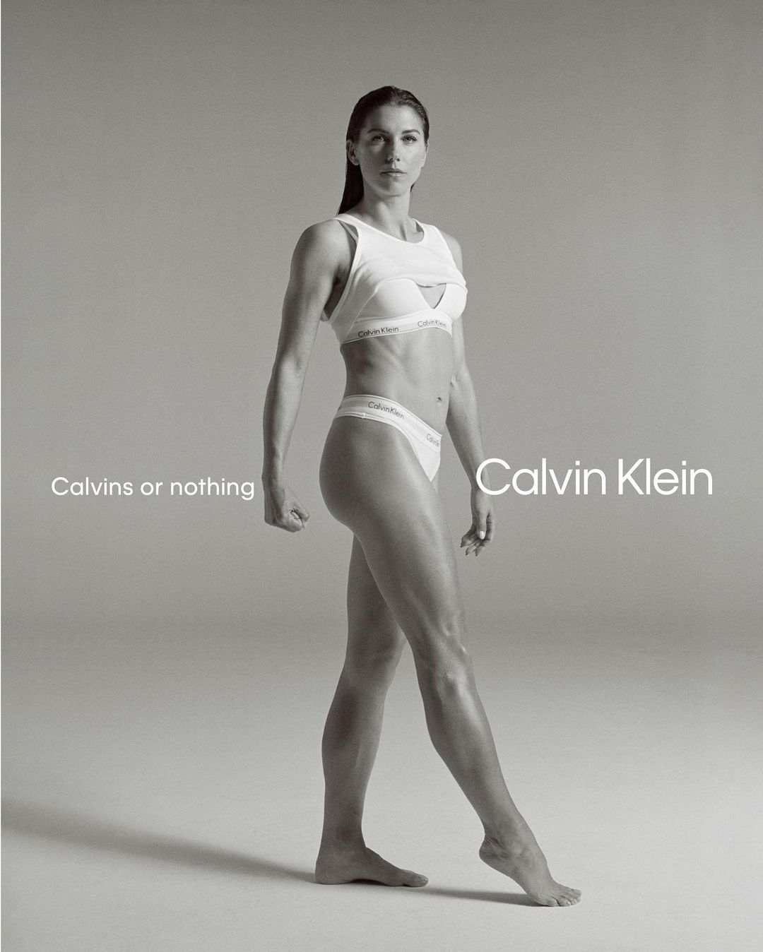 Alex Morgan standing against a seamless grey backdrop wearing white Calvin Klein athletic underwear and a matching sports bra.
