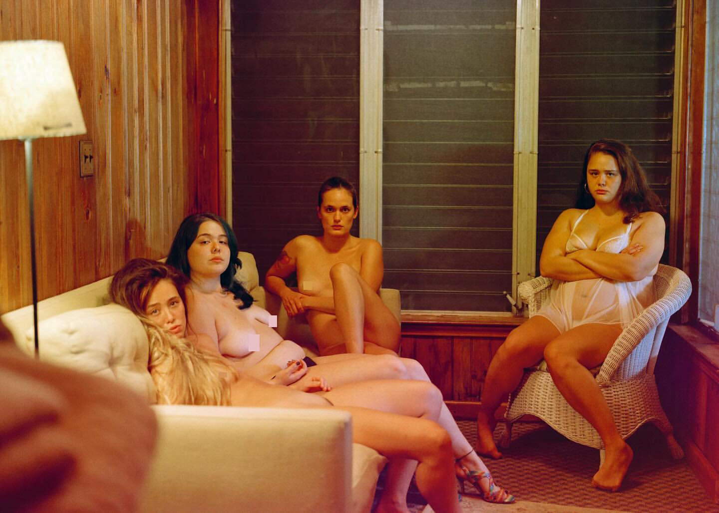 Four women pose intimately indoors; one seated in white lingerie with arms crossed, others reclining semi-nude on a light couch.
