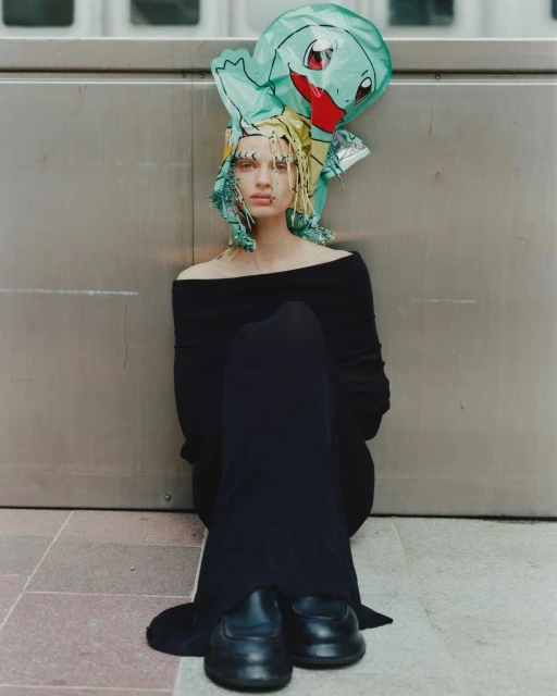 Woman in black off-shoulder top sits with a Squirtle balloon on her head in an urban setting, exuding unique style and modern fashion.